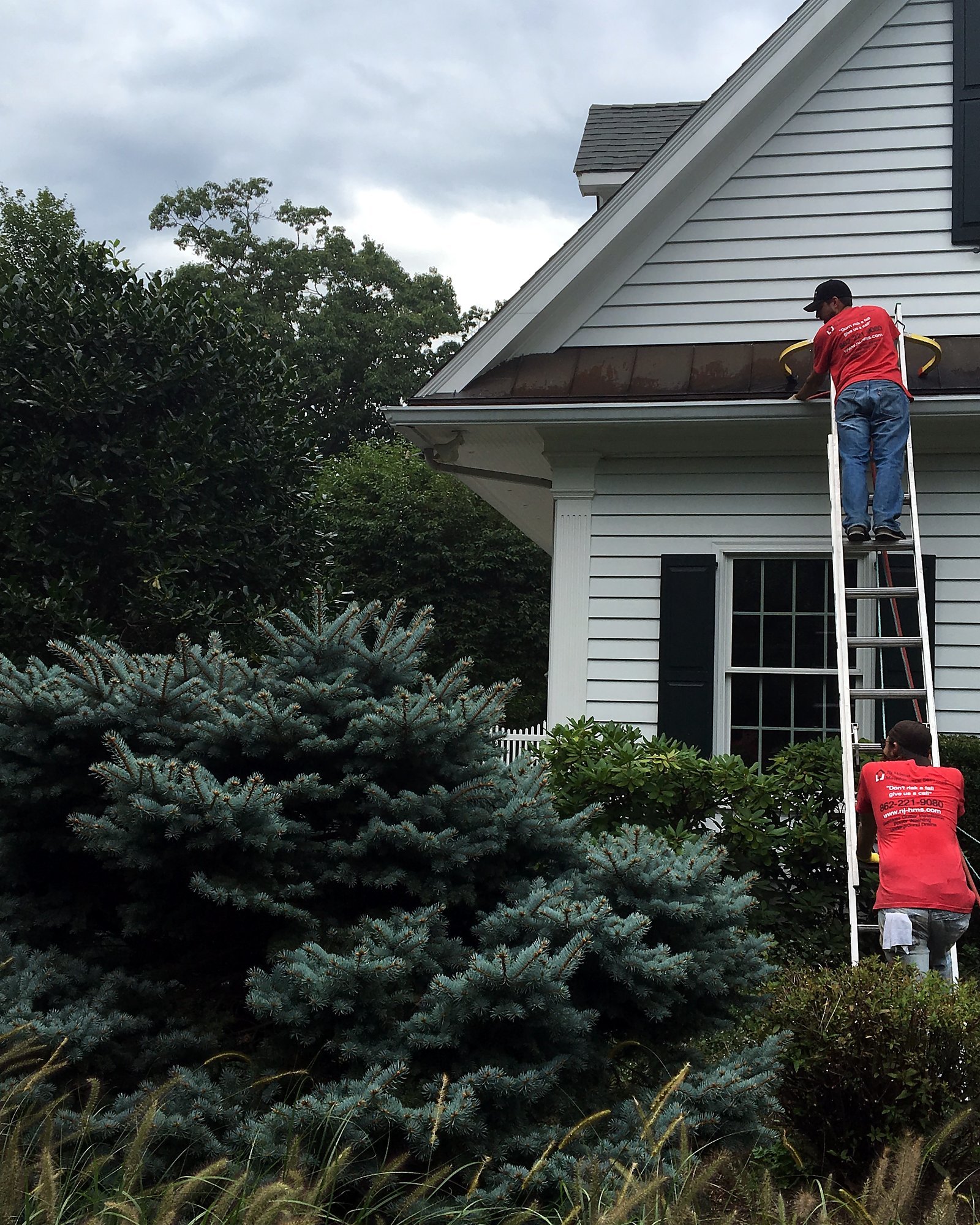 Two NJHMS technicians on a ladder cleaning gutters at a home in Franklin Lakes, NJ