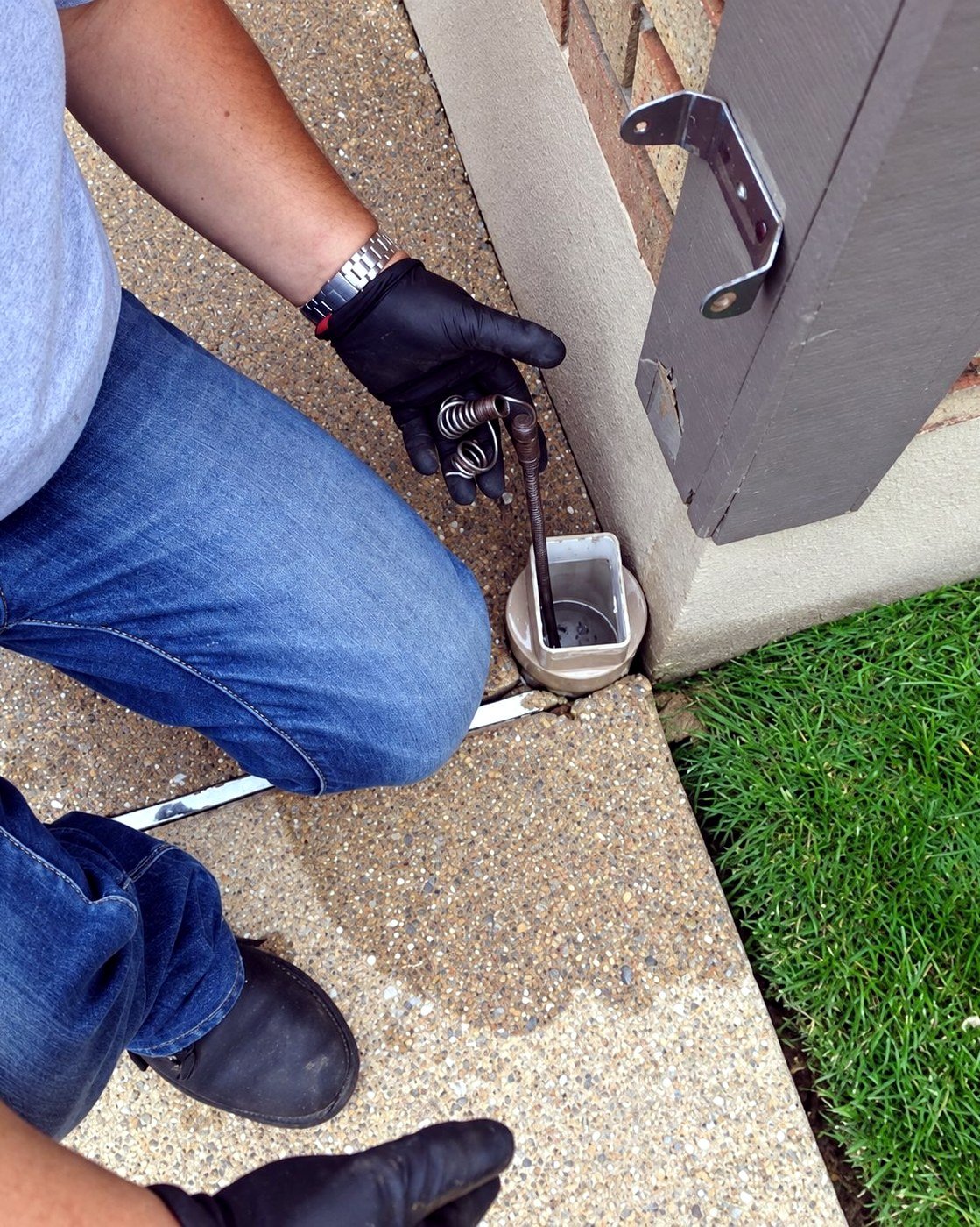 Technician using a hand auger to clear debris from a downspout drain connection at the base of a home
